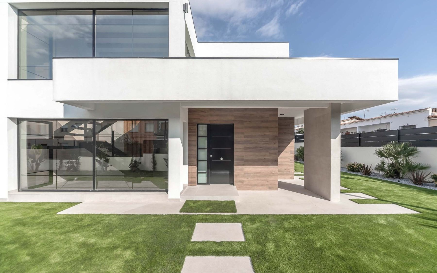 Casa modular de dos plantas con volúmenes blancos, entrada revestida en tono madera y jardín de césped en Masquefa
